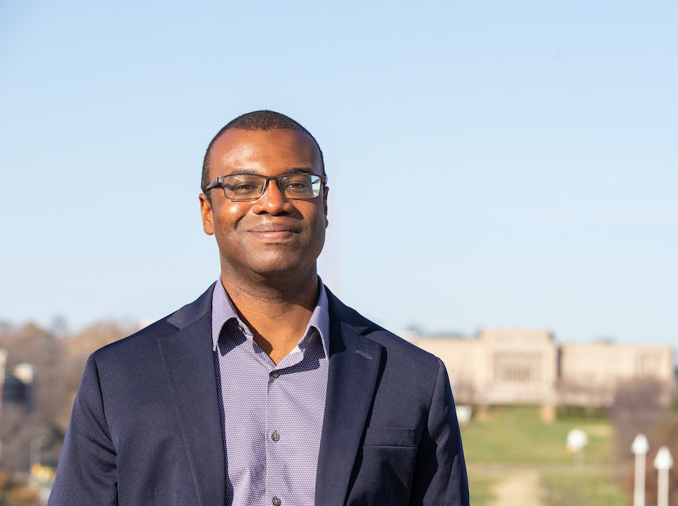 Image of Black man wearing a navy blue blazer and purple shirt; image of sky and museum in the distance background
